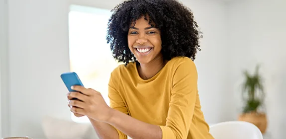 Jonge vrouw van kleur met een smartphone in haar hand kijkt lachend in de camera.